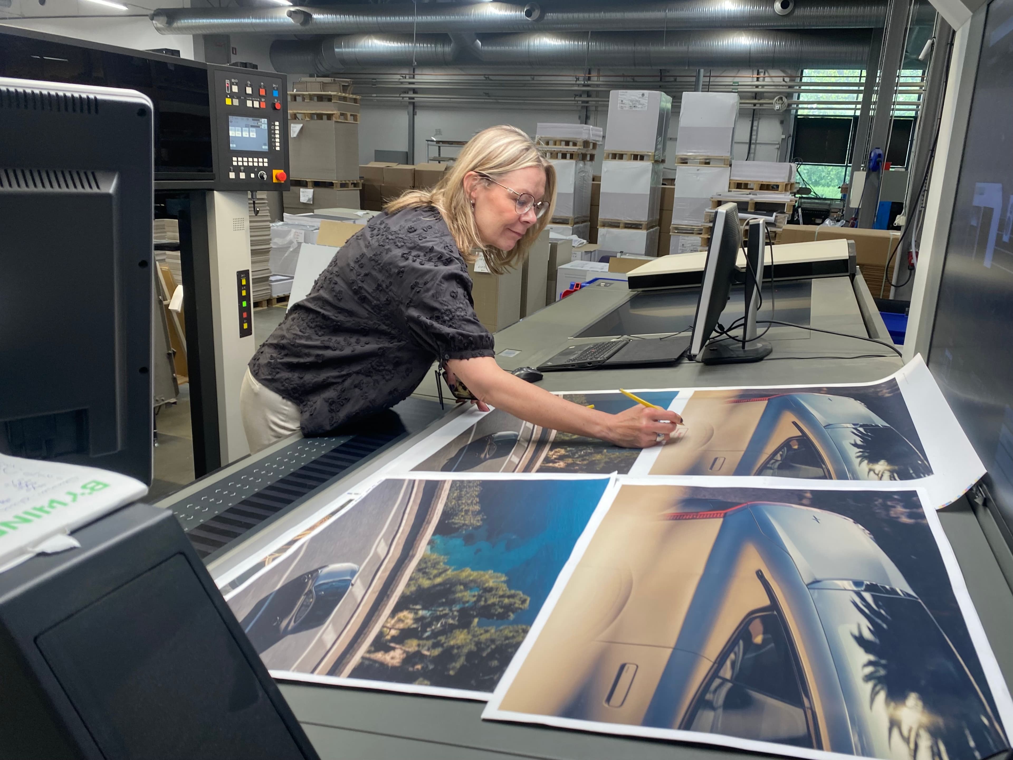 Print production specialist reviewing large-format automotive prints in a professional printing facility, showcasing advanced commercial printing and color management capabilities. The image highlights high-resolution image output, precision quality control, and state-of-the-art print technology—ideal for marketing materials, retail graphics, packaging prototypes, and premium brand campaigns. Perfect representation of end-to-end print and packaging solutions, from prepress and proofing to large-scale production.