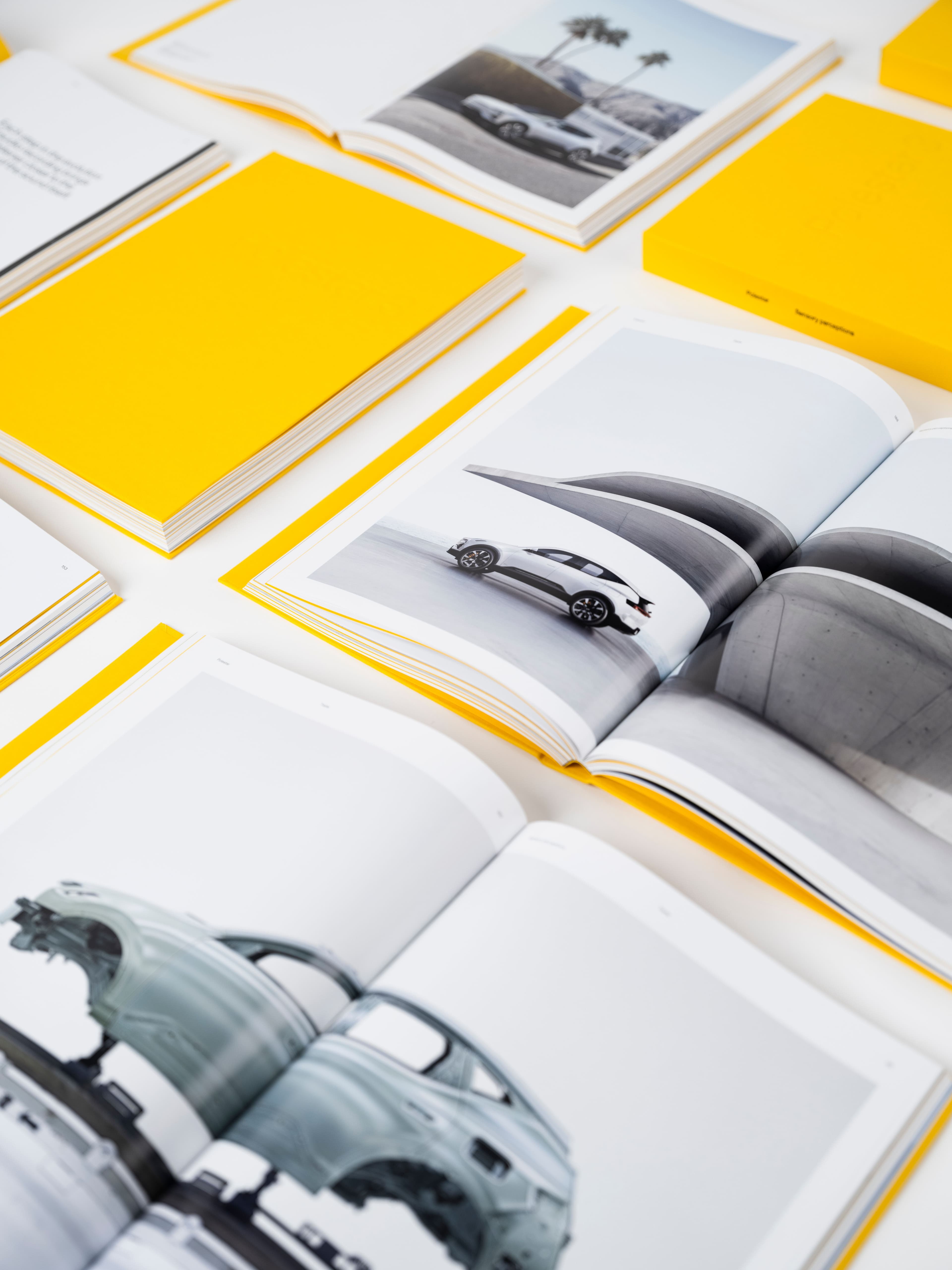 En modern layout för en elektrisk fordonskatalog med premiumfotografi av bilar, tryckt i högkvalitativa magasin med djärva gula omslag. Bilden visar både öppna och stängda redaktionella uppslag med eleganta sportbilar och detaljerade fordonsbilder, arrangerade i en ren, minimalistisk komposition mot vit bakgrund. En idealisk presentation av lyxigt fordonsvarumärke, printdesign och samtida boklayout för inspiration.